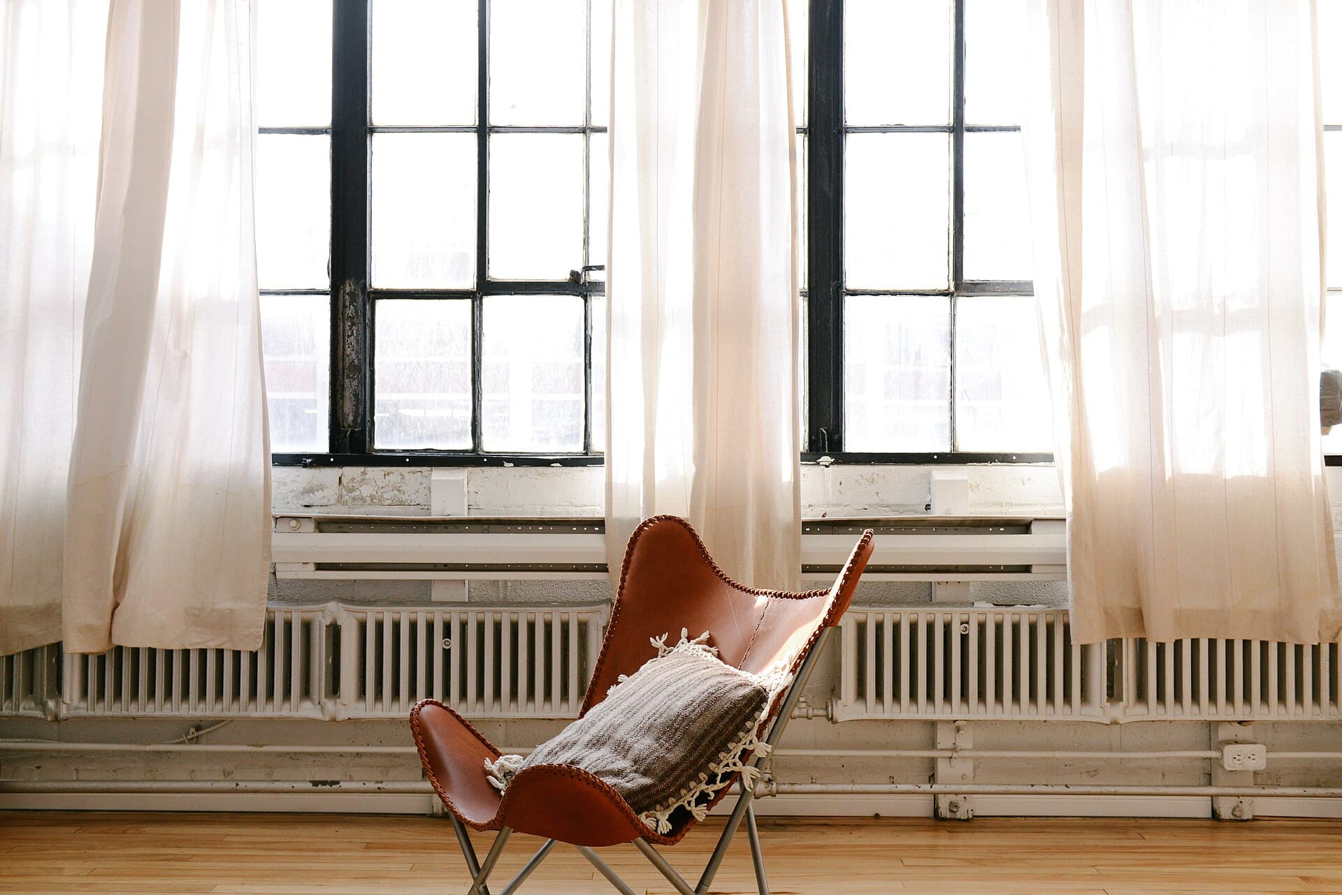 A leather sling chair in front of tall windows hung with sheer curtains
